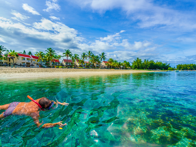 Berrak bir denizden çekilmiş şnorkel ile dalış yapan turist ve uzaktan görünen sahilde otel odaları