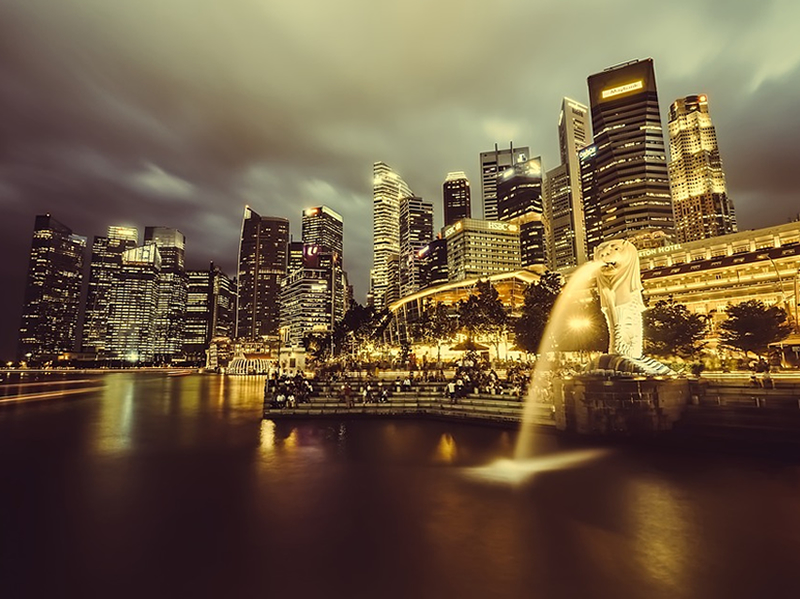 Gece sarı ışıklar ile aydınlatılmış nehir kenarındaki gökdelenler ve ağzından su fışkırtan merlion anıtı, Singapur